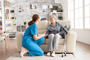 A female healthcare worker in scrubs talking with an elderly woman in her home.